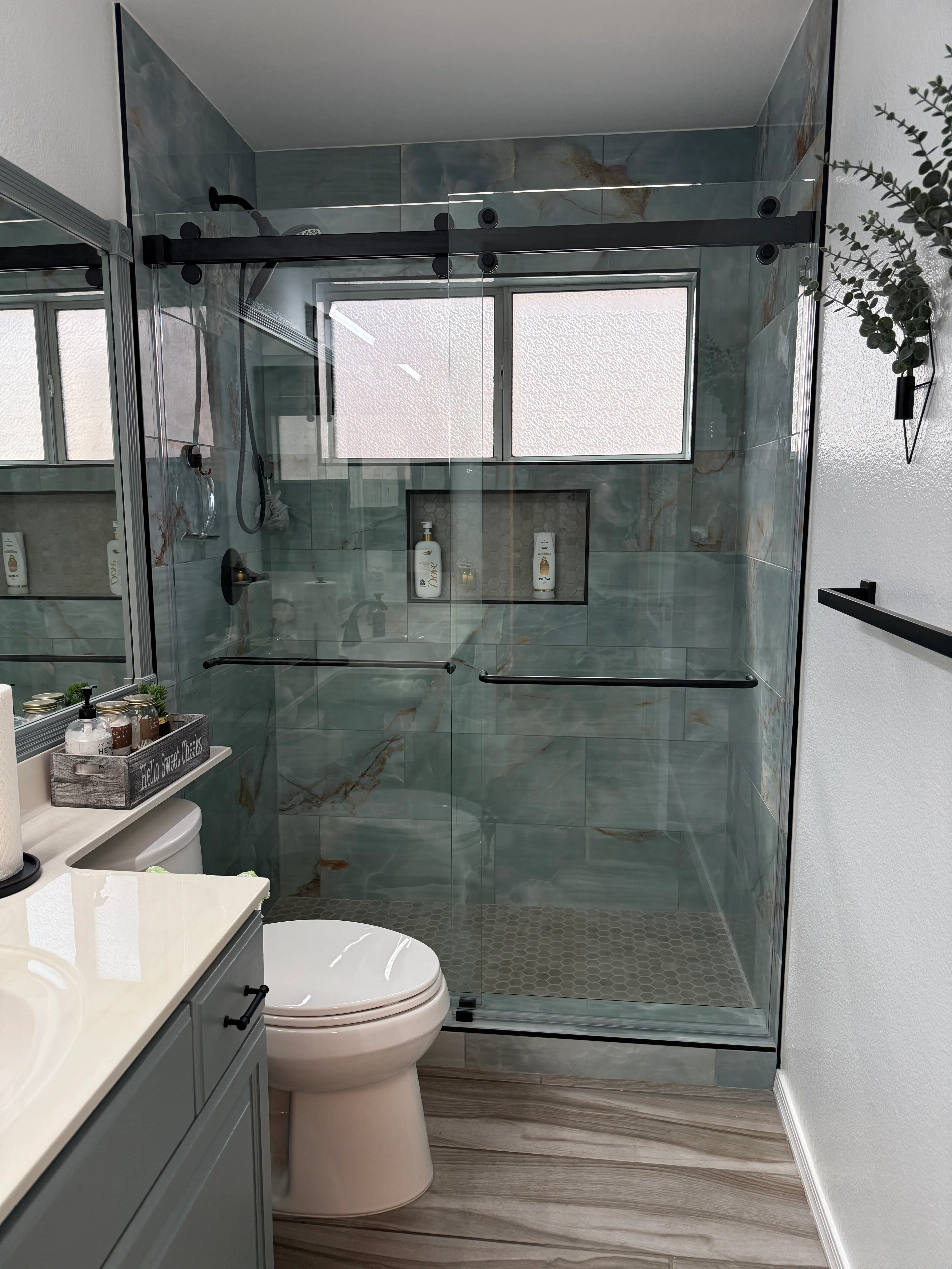 Frameless glass shower enclosure with gray tile walls, white vanity, and wood-look flooring